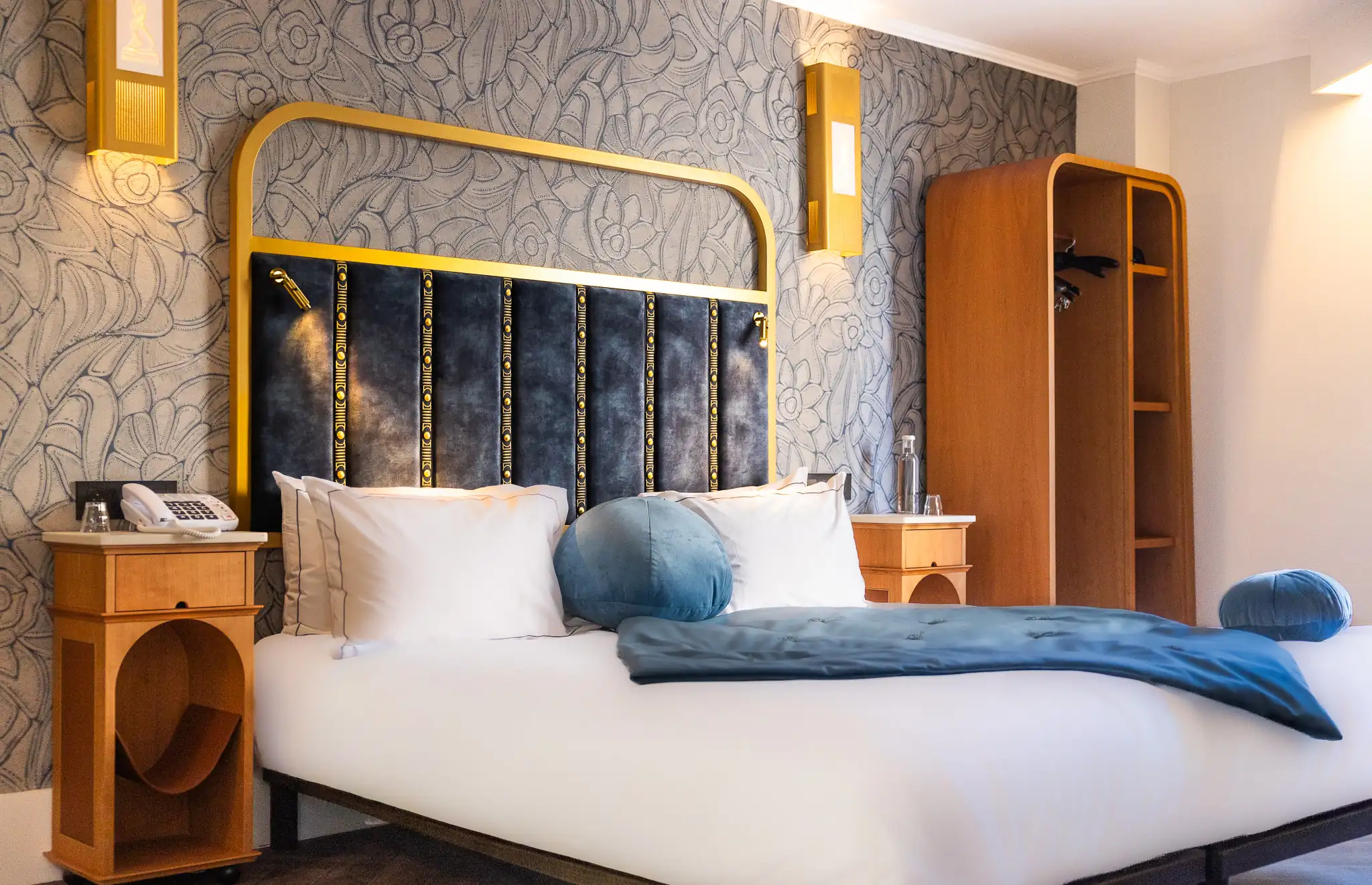 Chambre avec un lit double comportant une tête de lit bleue à cadres dorés, des coussins blancs et ronds bleus, une couverture bleue, deux tables de nuit en bois clair avec lampe murale dorée et un grand placard ouvert en bois à droite.