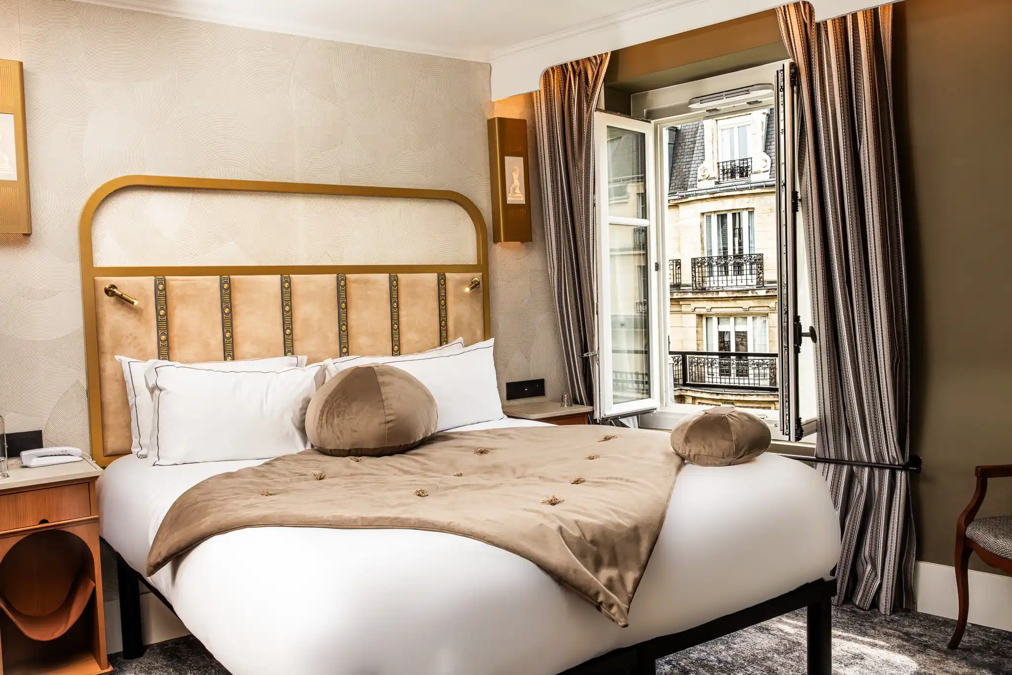 Chambre élégante avec lit double à tête de lit en velours beige bordé d'or, couvertures et coussins taupe, fenêtre ouverte donnant sur des bâtiments parisiens en pierre avec balcons en fer forgé, rideaux rayés et mobilier en bois clair.