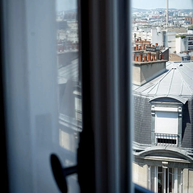 Vue sur les toits d'un bâtiment parisien avec des cheminées et une fenêtre à volets fermés, prise depuis une fenêtre intérieure floue.