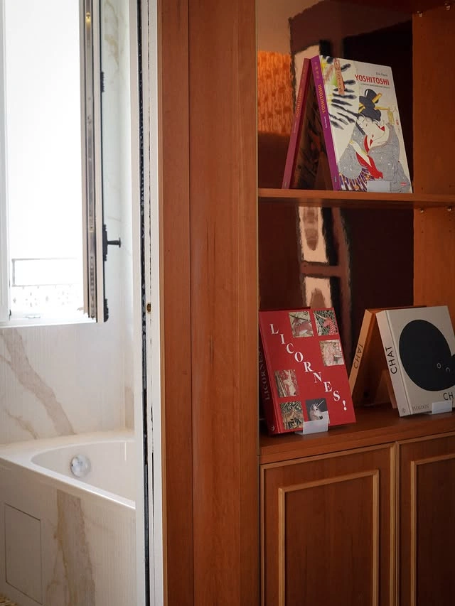 Bibliothèque en bois avec trois livres exposés devant une baignoire blanche visible par une fenêtre ouverte dans une salle de bain.