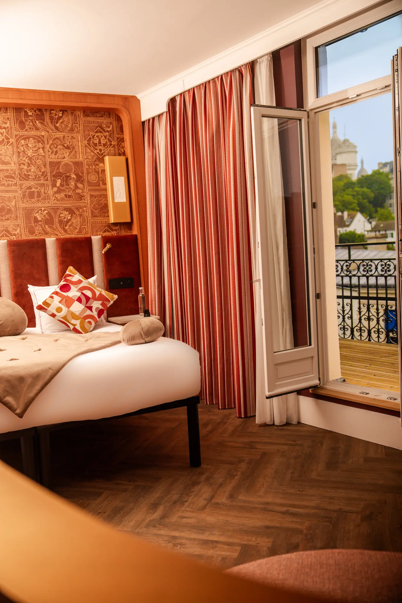 Chambre à coucher avec lit double orné de coussins colorés, tête de lit en bois sculpté, rideaux rayés rouge et blanc, et porte-fenêtre ouverte donnant sur un balcon en fer forgé avec vue partielle sur un bâtiment historique et des arbres.