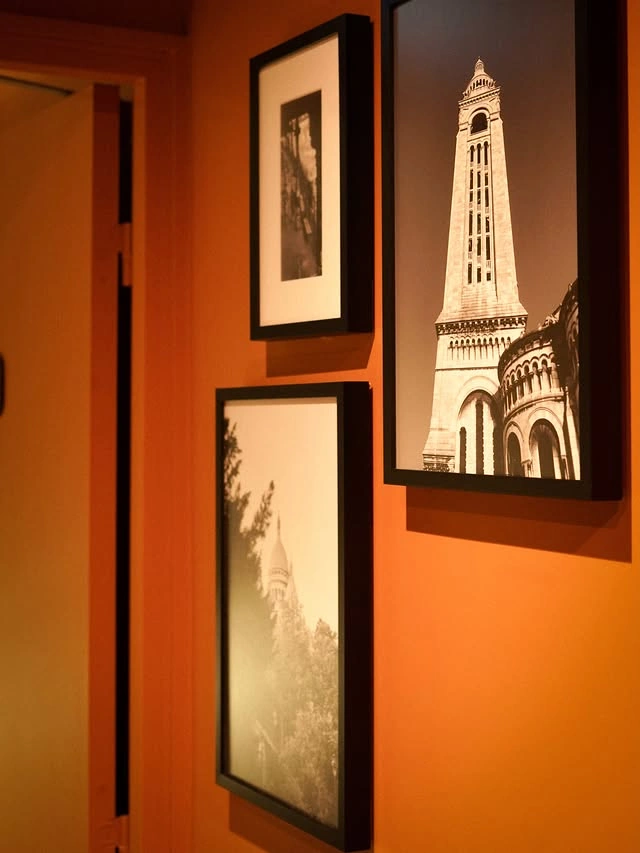 Trois cadres noirs accrochés sur un mur orange, contenant des photos en noir et blanc d'architectures, avec une porte partiellement visible à gauche.