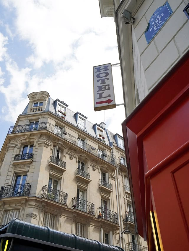 Façade d'un immeuble haussmannien avec balcons en fer forgé sous un ciel partiellement nuageux, à côté d'un panneau vertical indiquant un hôtel avec une flèche rouge pointant vers la droite, et une porte rouge au premier plan.