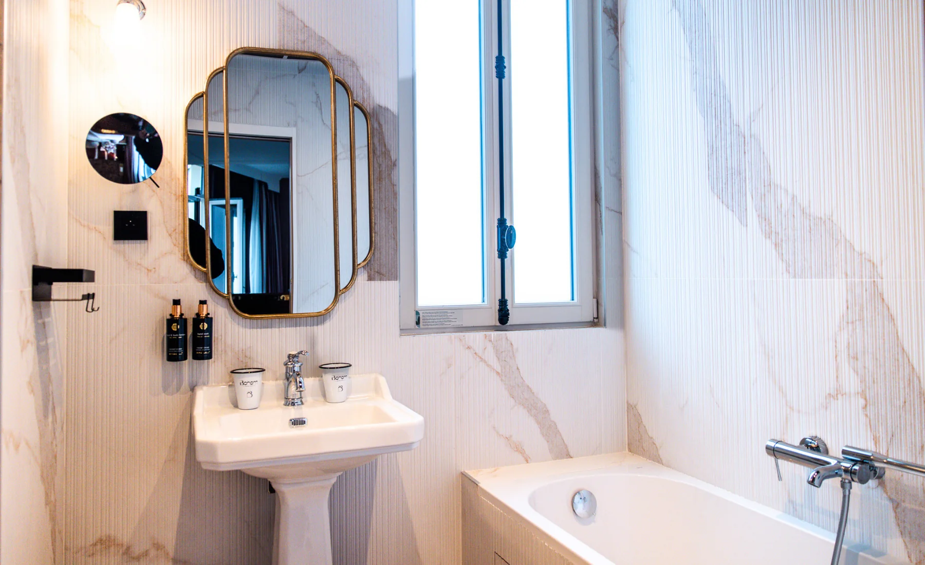 Salle de bain élégante avec lavabo blanc sur pied contenant deux tasses, miroir mural doré à bord arrondi, deux distributeurs de savon noirs fixés au mur, fenêtre centrale avec volet bleu et baignoire blanche équipée d'un robinet chromé.