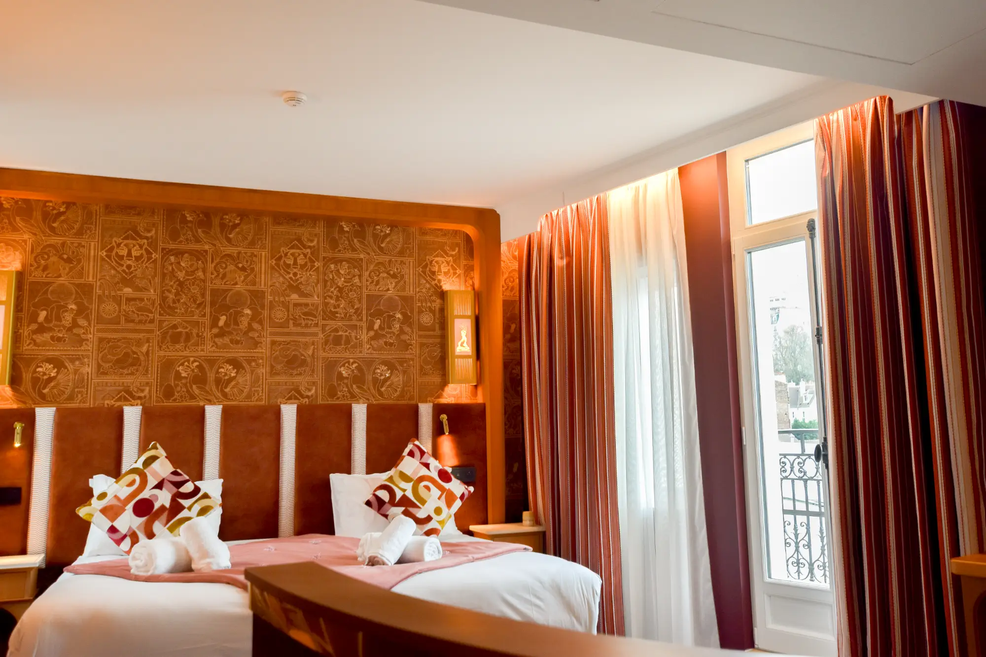 Chambre d'hôtel avec un grand lit double décoré de coussins colorés et de serviettes roulées, mur avec papiers peints ornés, une grande fenêtre avec rideaux rayés rouges et blancs laissant entrer la lumière naturelle.