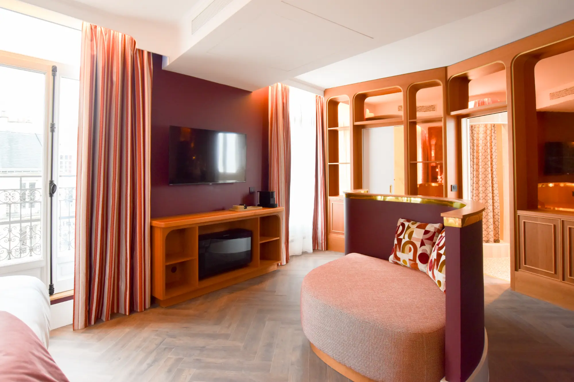 Chambre d'hôtel moderne avec mur violet foncé, télévision murale, meubles en bois clair, fauteuil arrondi avec coussins colorés, sol parquet et grandes fenêtres avec rideaux rayés.