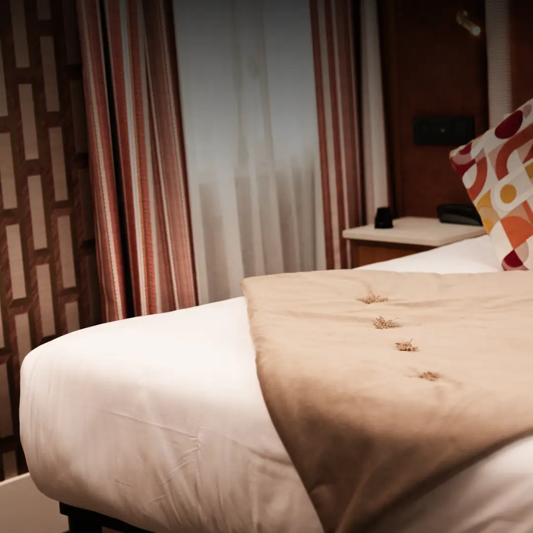 Cama de casal com colcha bege decorada com botões, travesseiro com estampas coloridas, abajur, cortinas listradas em vermelho e branco e parede com papel de parede de padrões geométricos.