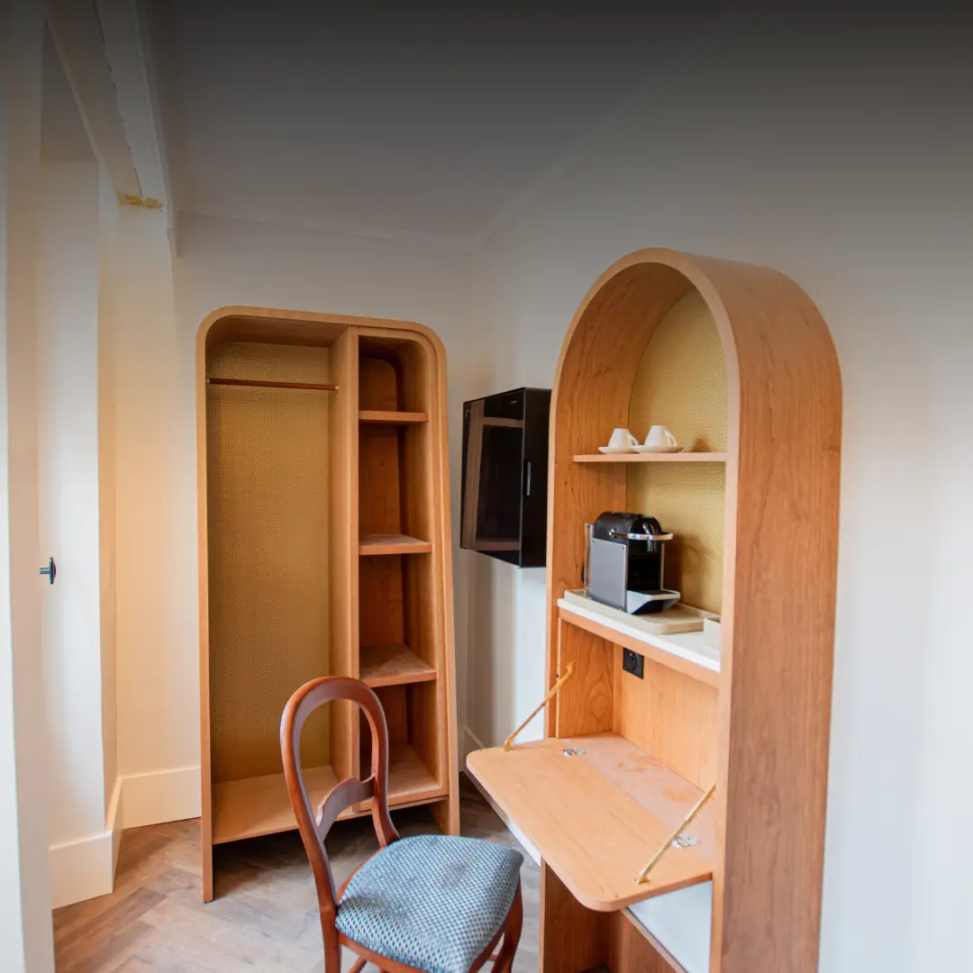 Coin bureau avec petit meuble en bois cintré abritant une machine à café avec deux tasses sur une étagère, chaise en bois avec siège rembourré à motif gris, et armoire ouverte avec étagères et tringle à gauche dans une pièce aux murs blancs.