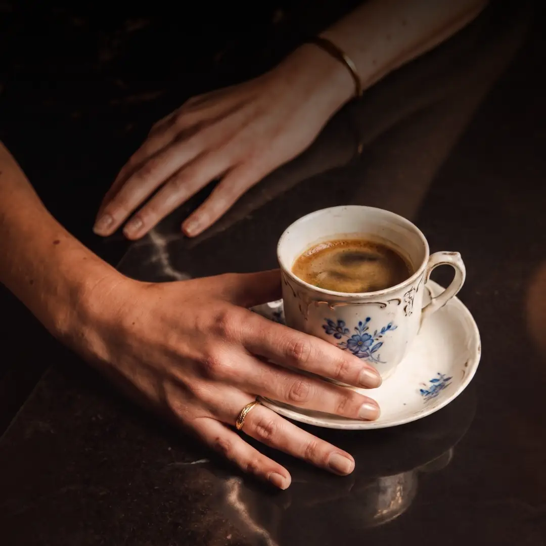 Mains féminines posées sur une surface sombre, l'une tenant une tasse de café dans une tasse blanche avec des motifs floraux bleus sur une soucoupe assortie.