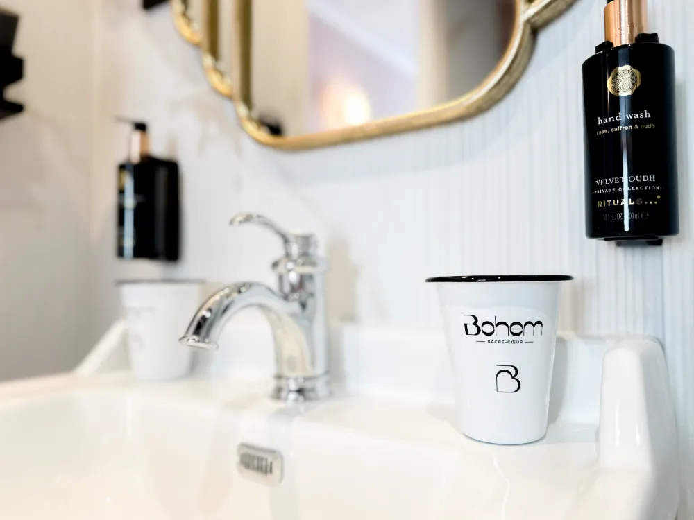 Évier blanc avec robinet chromé dans une salle de bain moderne, un gobelet blanc avec l'inscription Bohem Sacré-Cœur posé sur le bord de l'évier, à côté d'un distributeur mural noir de savon liquide Rituals Velvet Oudh.
