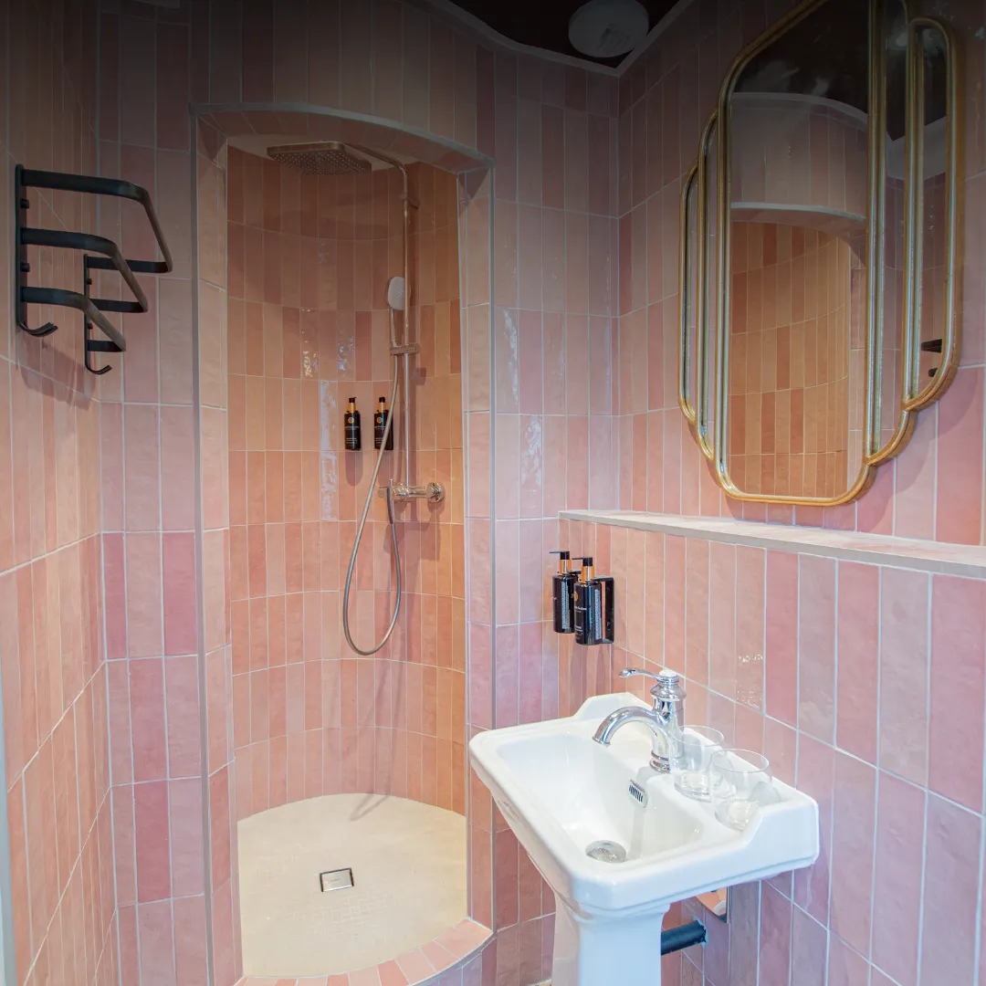 Salle de bain avec douche ouverte carrelée en rose, pomme de douche avec flexible, deux distributeurs de savon, lavabo blanc avec robinet chromé, deux gobelets transparents et grand miroir doré mural.