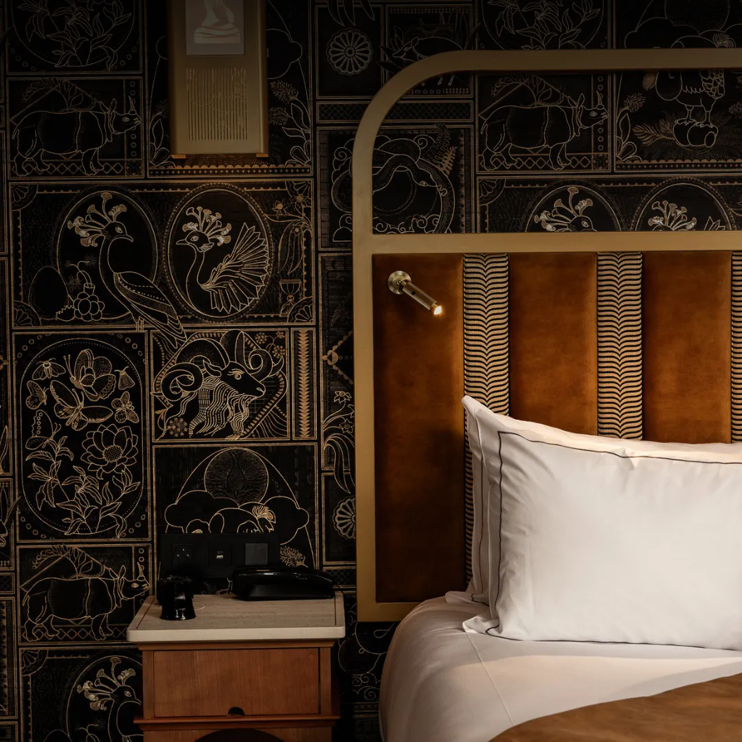Coin de chambre stylé avec tête de lit en velours camel et beige, lampe de lecture murale, oreiller blanc et table de chevet en bois clair devant un mur décoré de papier peint noir orné de motifs animaliers dorés.