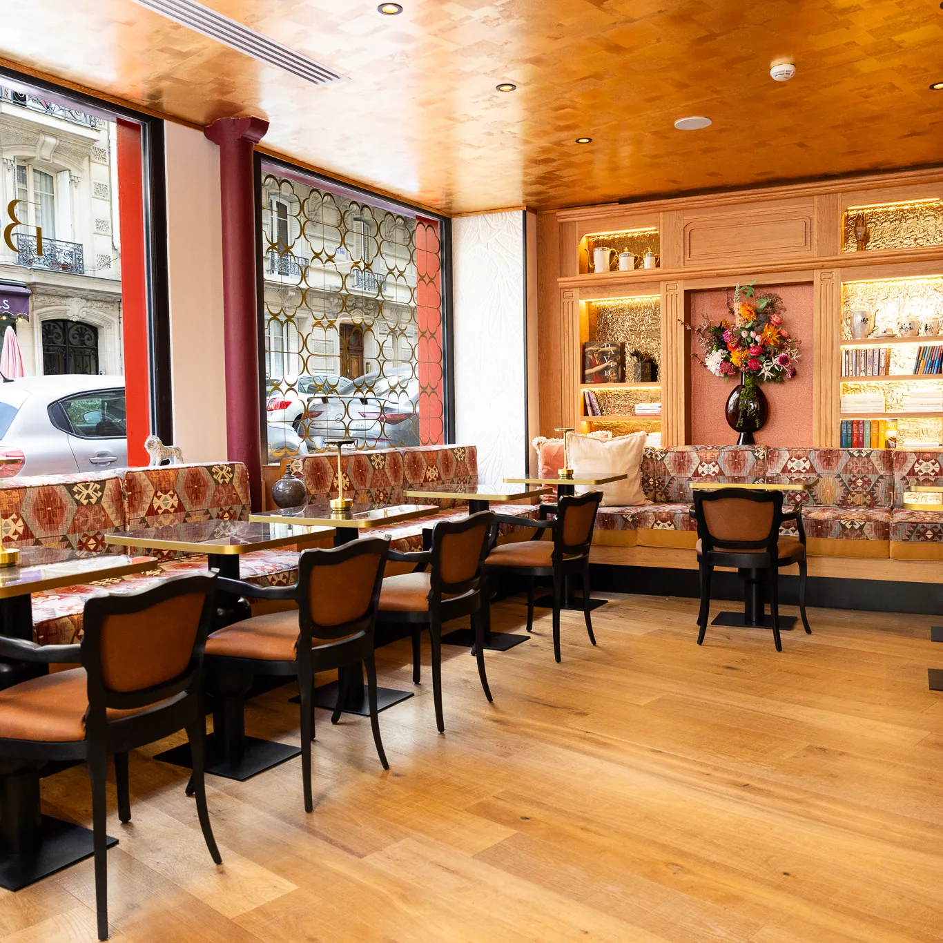Intérieur lumineux et élégant d'un café avec de grandes fenêtres, banquettes à motifs, chaises en bois, tables en marbre et étagères décorées de livres et d'un vase de fleurs.