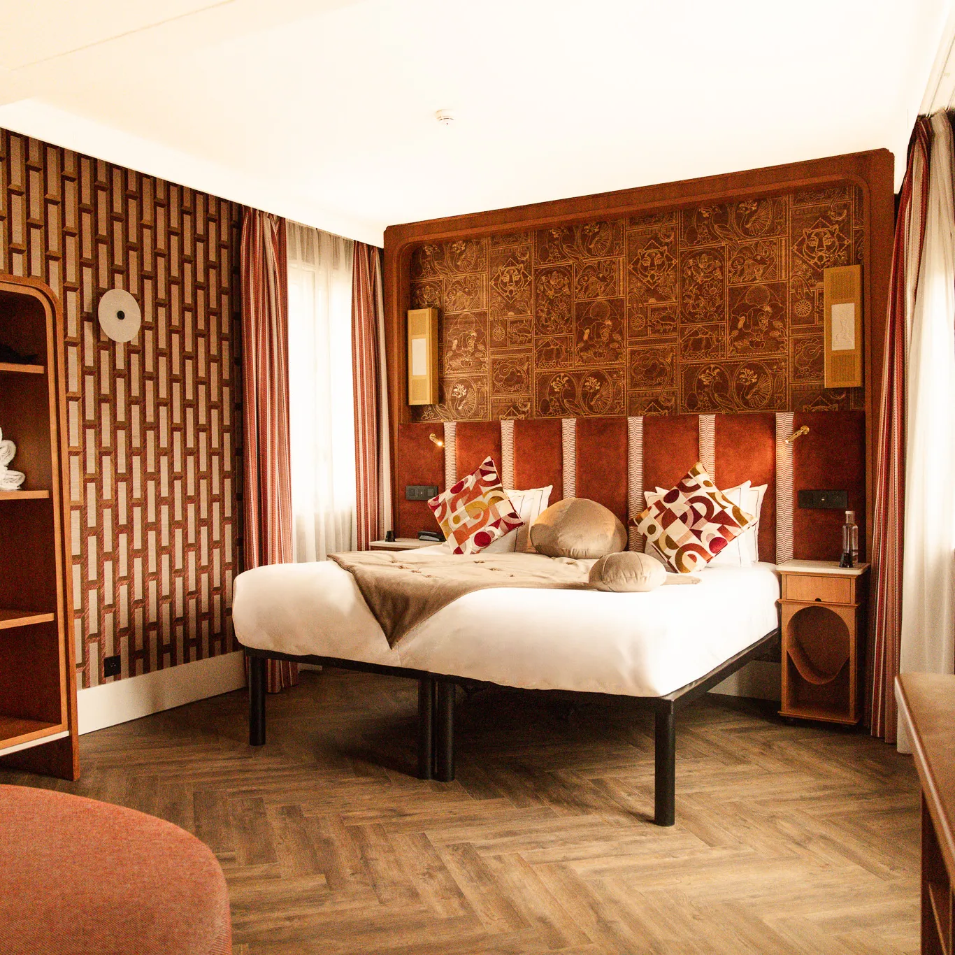 Chambre avec un grand lit double, oreillers décoratifs aux motifs rouges et beiges, tête de lit rouge ornée de panneaux artistiques, murs avec papier peint géométrique marron, rideaux rayés, parquet en bois et petite table de chevet.