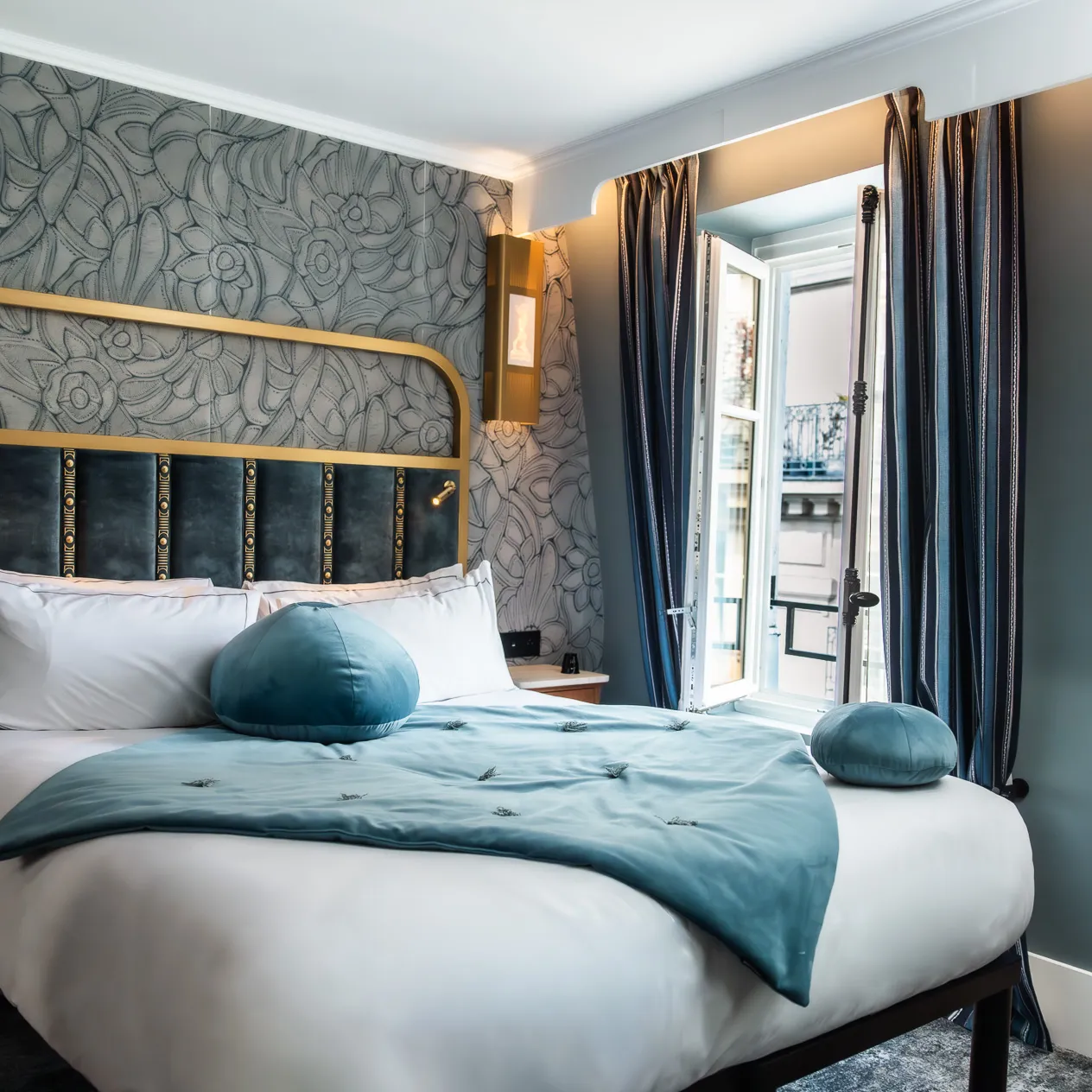 Chambre d'hôtel élégante avec un grand lit double portant une couette et deux coussins ronds bleu clair, une tête de lit en velours bleu marine avec cadre doré, un mur décoré d'un papier peint floral gris, une fenêtre ouverte encadrée de rideaux bleus foncés, et une lampe murale dorée.