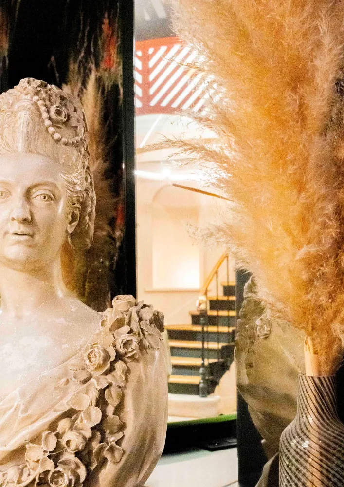 Buste sculpté d'une femme avec un haut orné de roses en relief, placé devant un miroir reflétant un escalier intérieur, avec un bouquet de plumeaux secs dans un vase à motifs sur le côté droit.