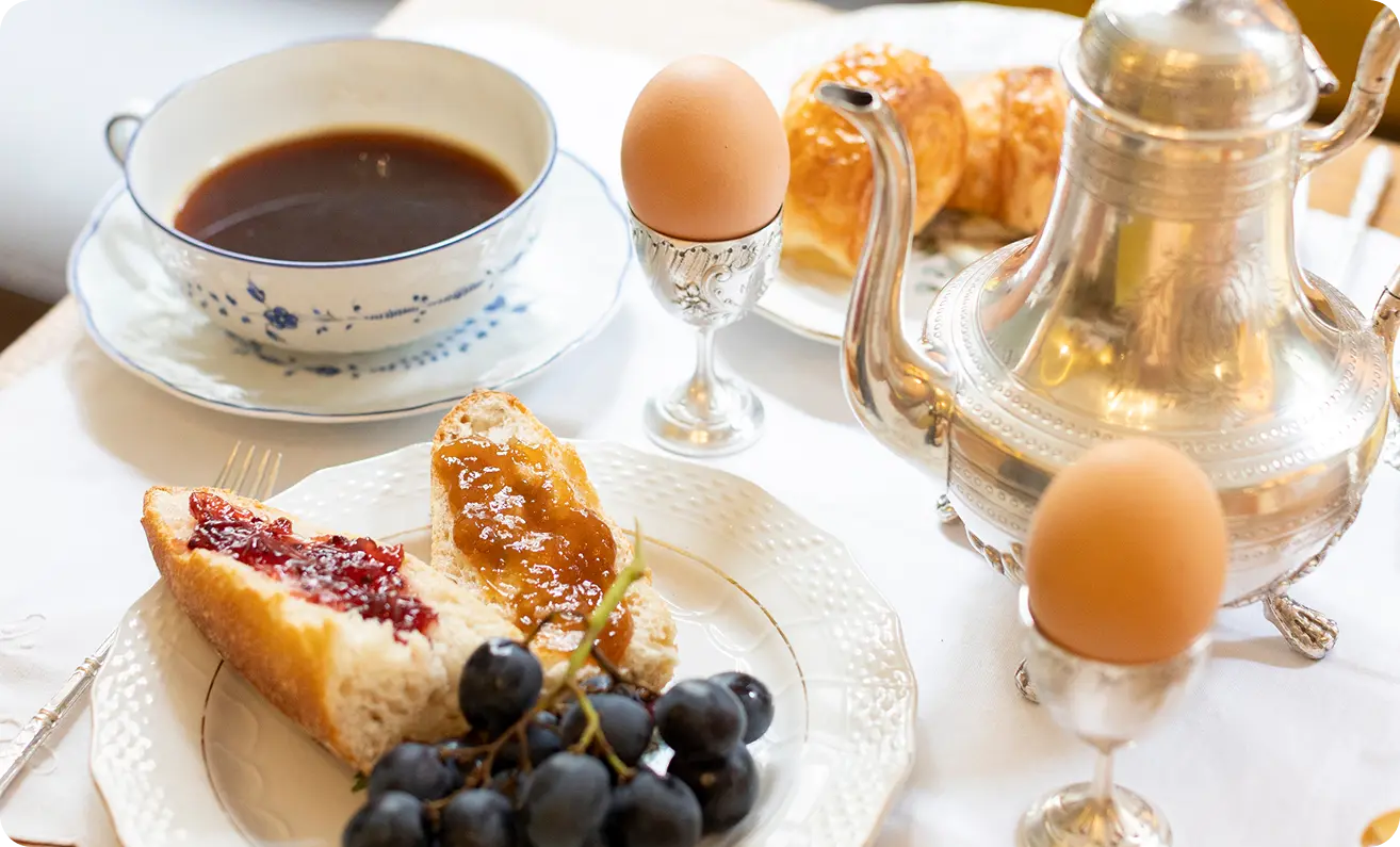 Allestimento per la colazione con tazza di caffè, teiera in argento, uova alla coque, pane con marmellata e uva su una tovaglia bianca.