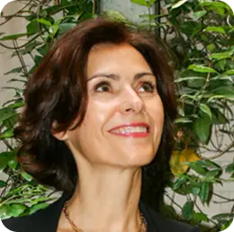 Woman with dark, wavy hair smiling and looking up, with green leafy plants in the background.
