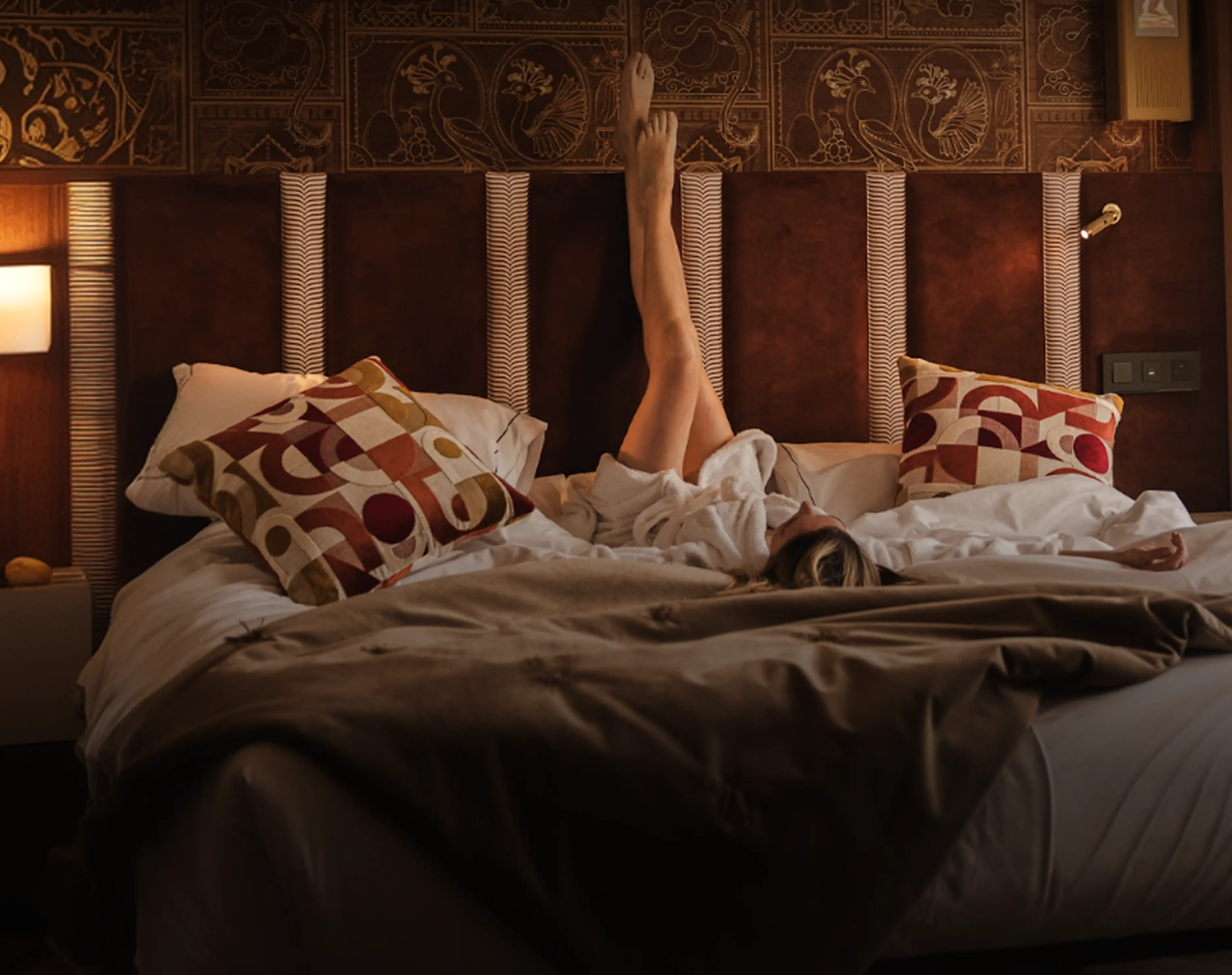 Personne allongée dans un lit avec une robe de chambre blanche et les jambes levées contre une tête de lit ornée de motifs artistiques, avec deux coussins colorés et une couverture marron sur le lit.