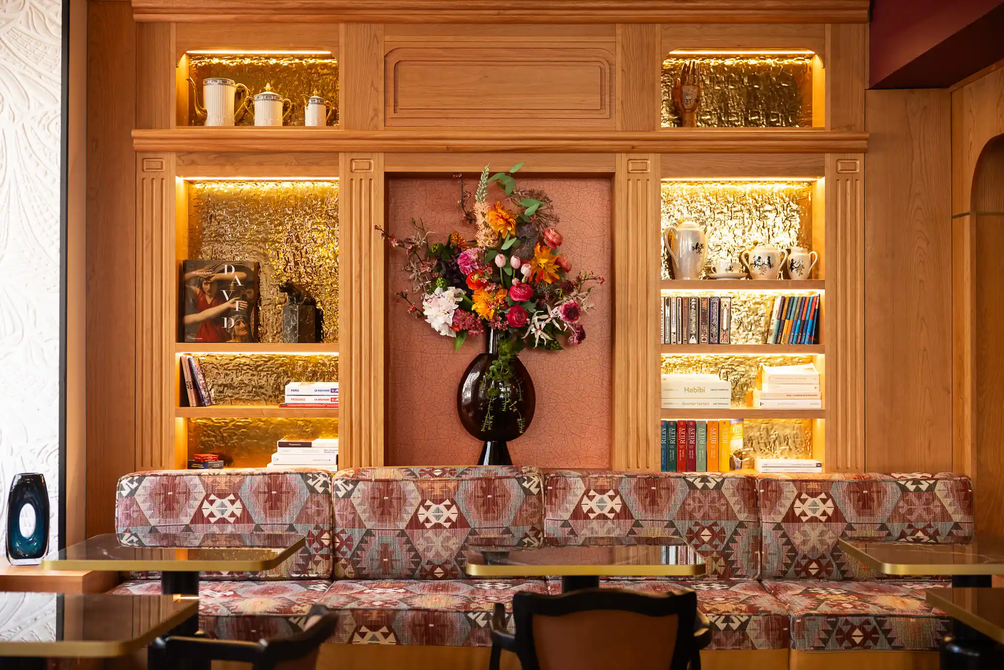 Coin salon avec une banquette à motifs géométriques rouges et beige devant une étagère en bois éclairée contenant des livres, de la vaisselle et un grand vase noir avec un bouquet de fleurs colorées.