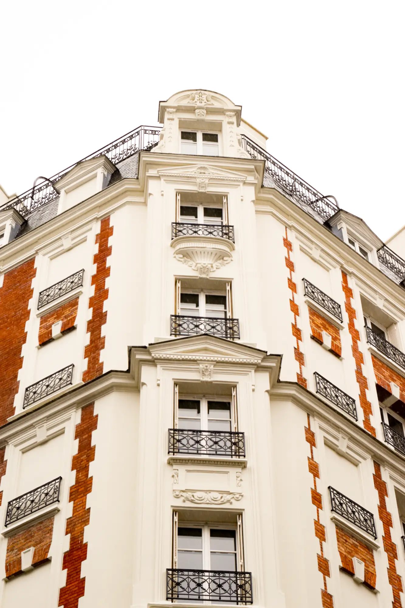 Façade d'un bâtiment urbain de style classique avec des murs blancs et des briques rouges, fenêtres à balcons en fer forgé, et un toit mansardé avec des lucarnes.