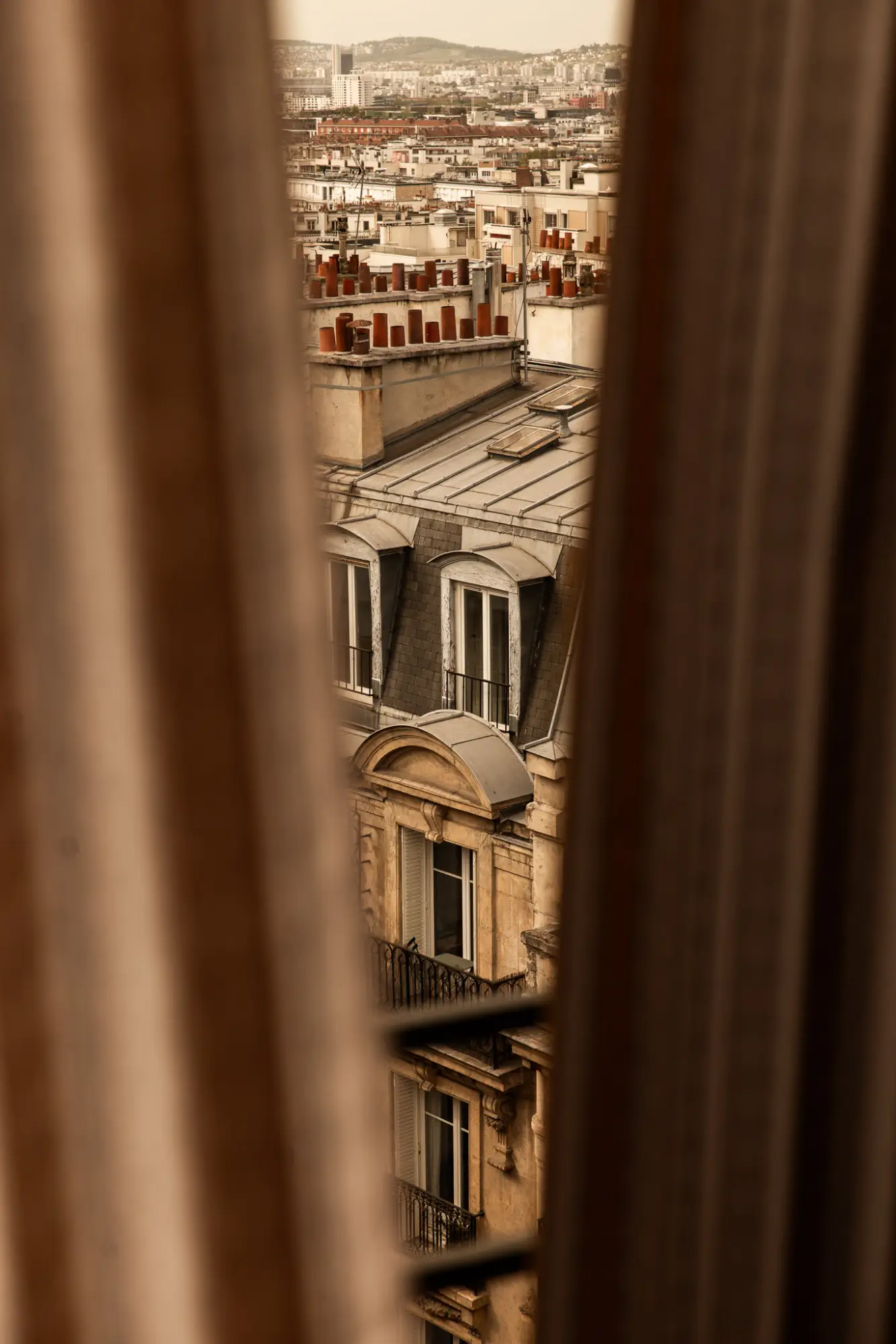 Vue urbaine d'immeubles parisiens à travers une fenêtre partiellement couverte par des rideaux, avec des toits en zinc et des cheminées en brique rouge.