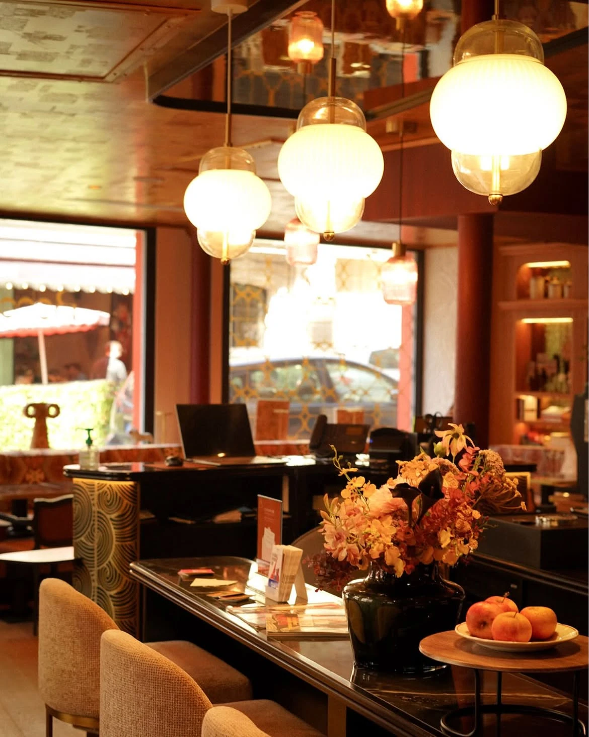 Intérieur d'un restaurant avec un comptoir en verre, un vase noir avec un bouquet de fleurs colorées, une assiette avec des pommes, et des lampes suspendues rondes éclairant chaleureusement la pièce.
