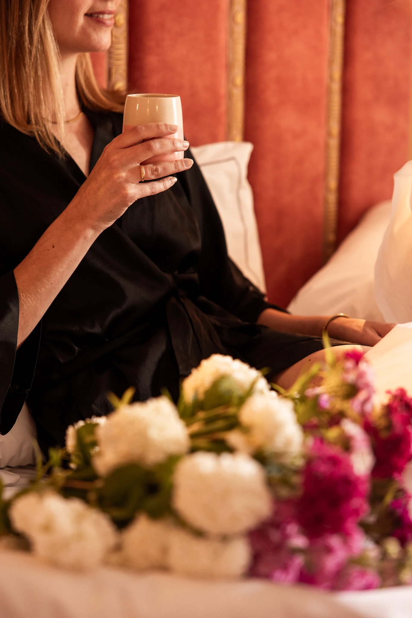 Une femme en robe noire tenant une tasse blanche et souriante, assise sur un lit avec une tête de lit rouge et des coussins blancs, un bouquet de fleurs blanches et roses au premier plan flou.
