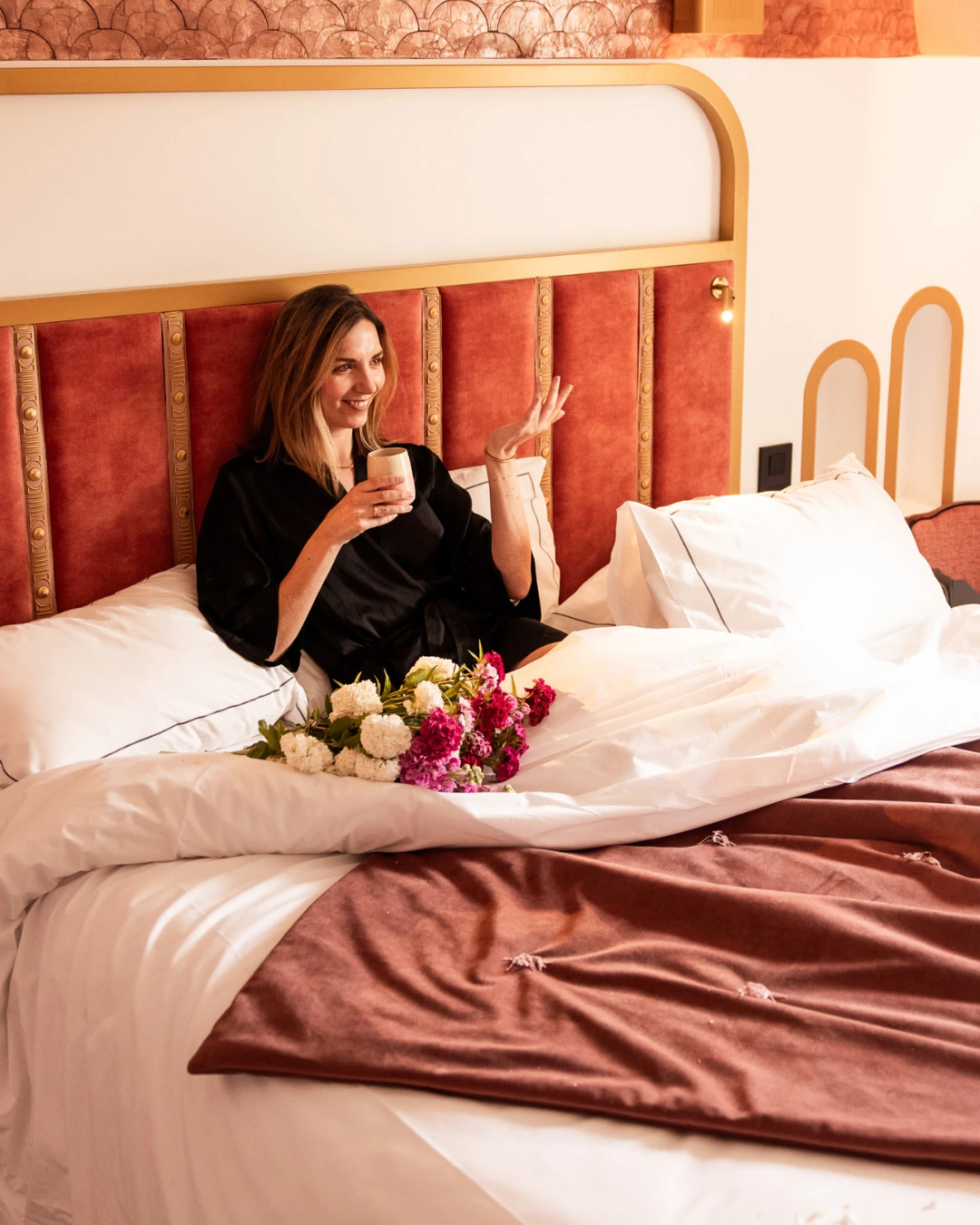 Une femme souriante en peignoir noir est assise dans un lit avec une tête de lit rouge et dorée, tenant une tasse, avec un bouquet de fleurs blanches et roses devant elle sur la literie blanche et une couverture marron.