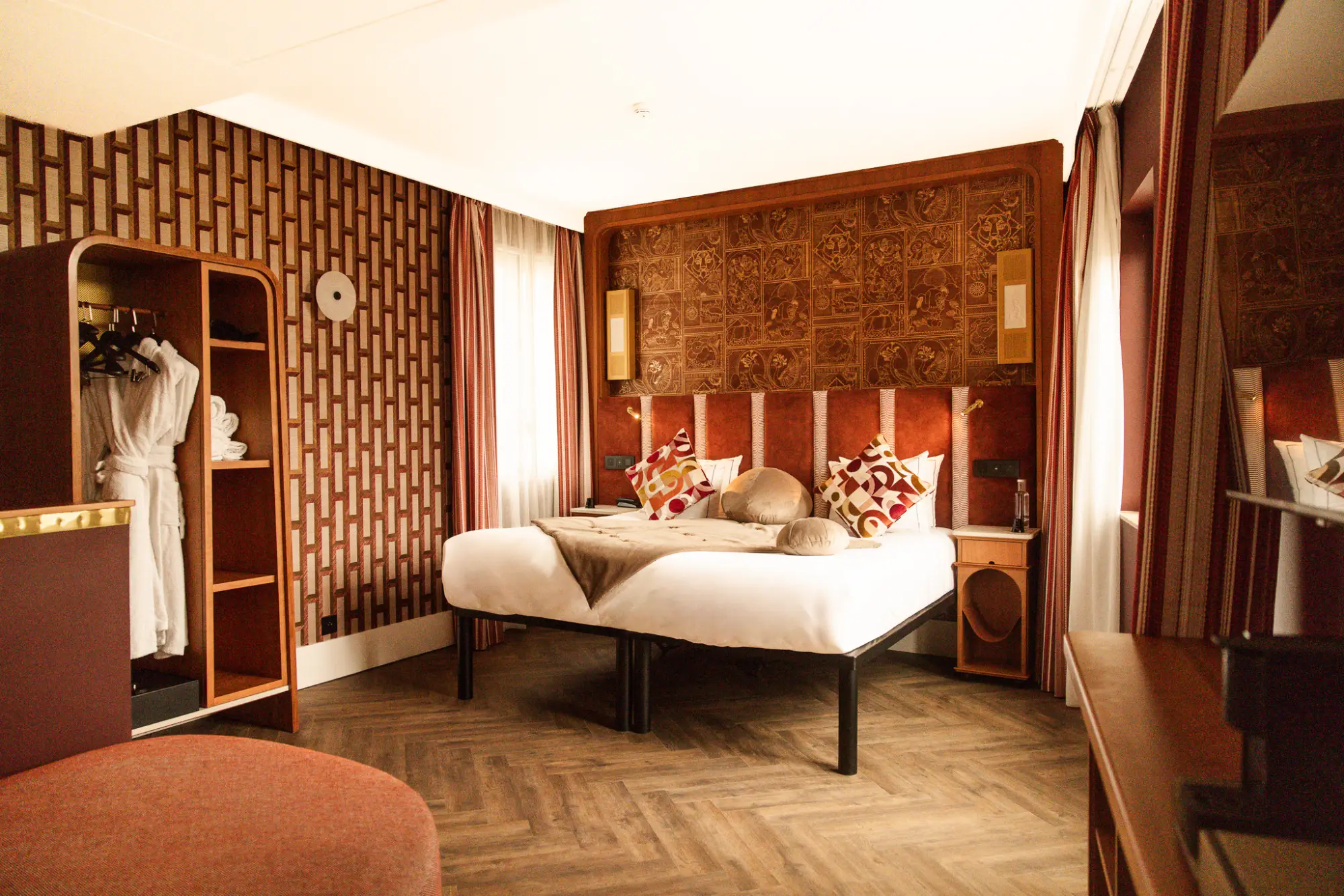Chambre d'hôtel élégante avec un grand lit double décoré de coussins colorés et de couvertures beige, un mur décoratif en bois sculpté derrière le lit, des rideaux rouges et un sol en bois clair à motif chevron.