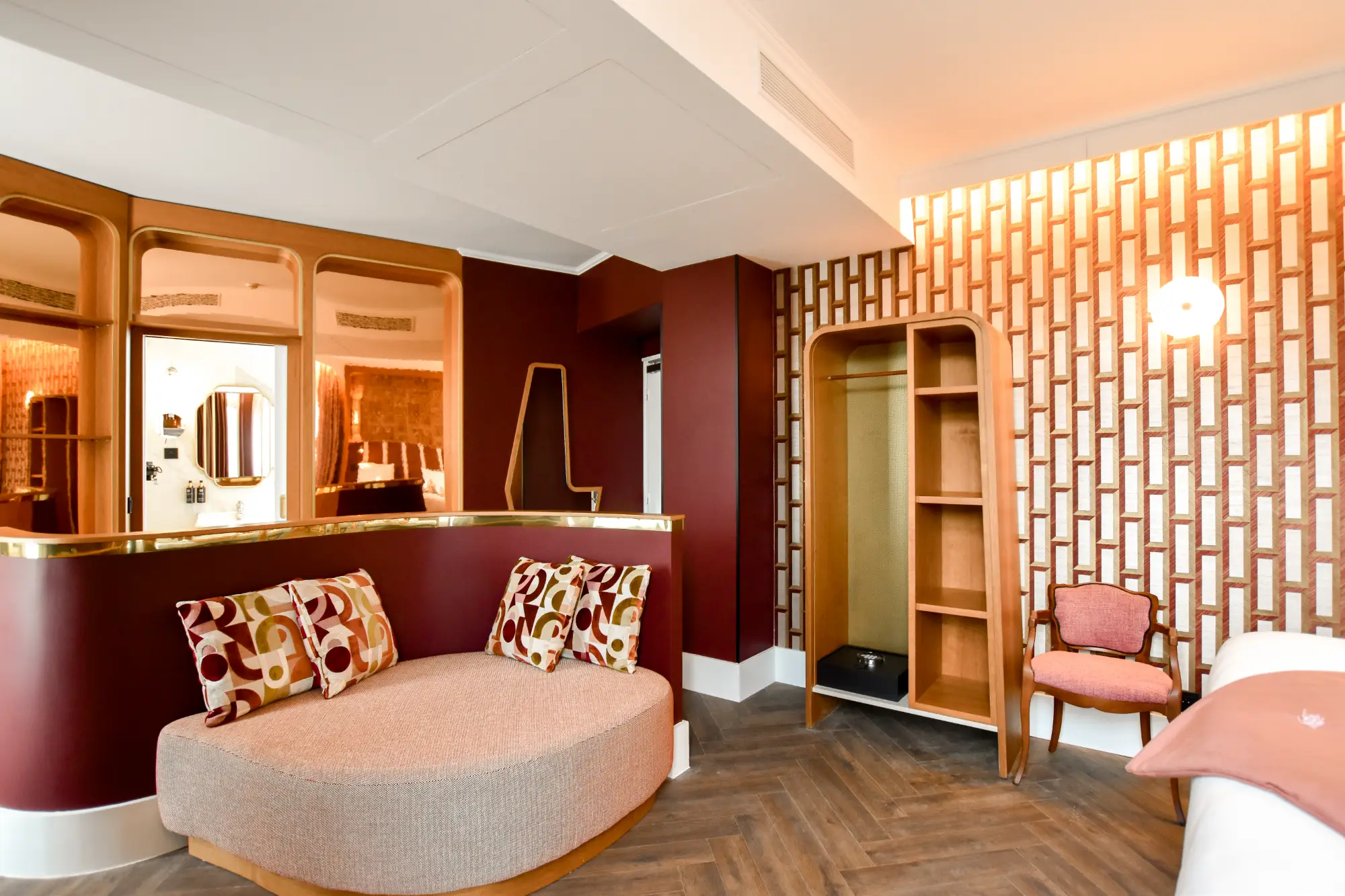 Intérieur d'une chambre d'hôtel avec un canapé arrondi beige orné de coussins aux motifs géométriques rouges, oranges et verts, un mur rouge foncé avec une séparation en verre miroirs, une armoire en bois ouverte avec étagères à côté d'un fauteuil rose et un mur avec un papier peint géométrique orange et blanc, éclairé par une applique murale ronde blanche.