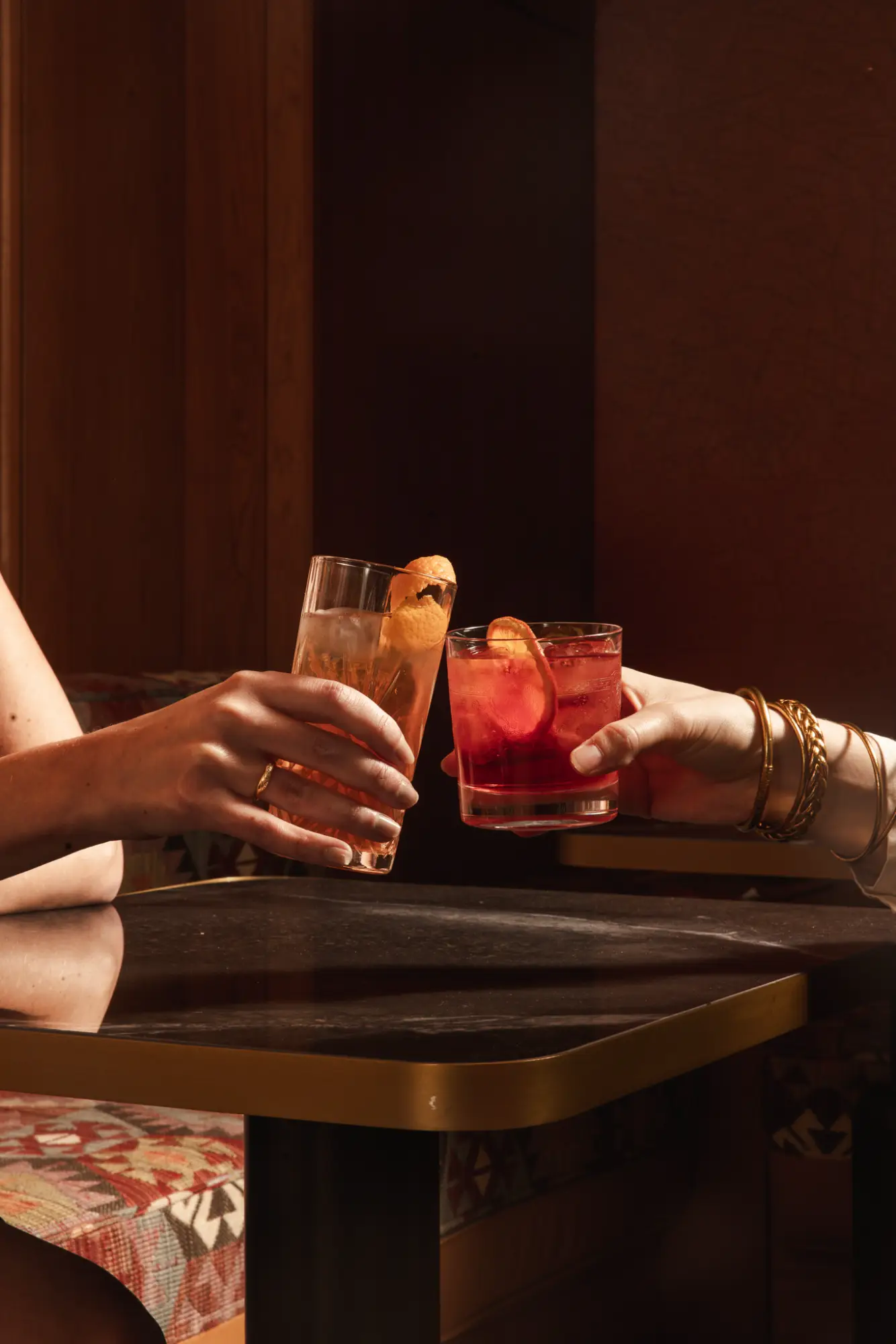 Deux mains tenant des verres à cocktail lèvent un toast au-dessus d'une table noire avec une bordure dorée, dans un cadre intimiste avec des sièges au tissu aux motifs colorés.