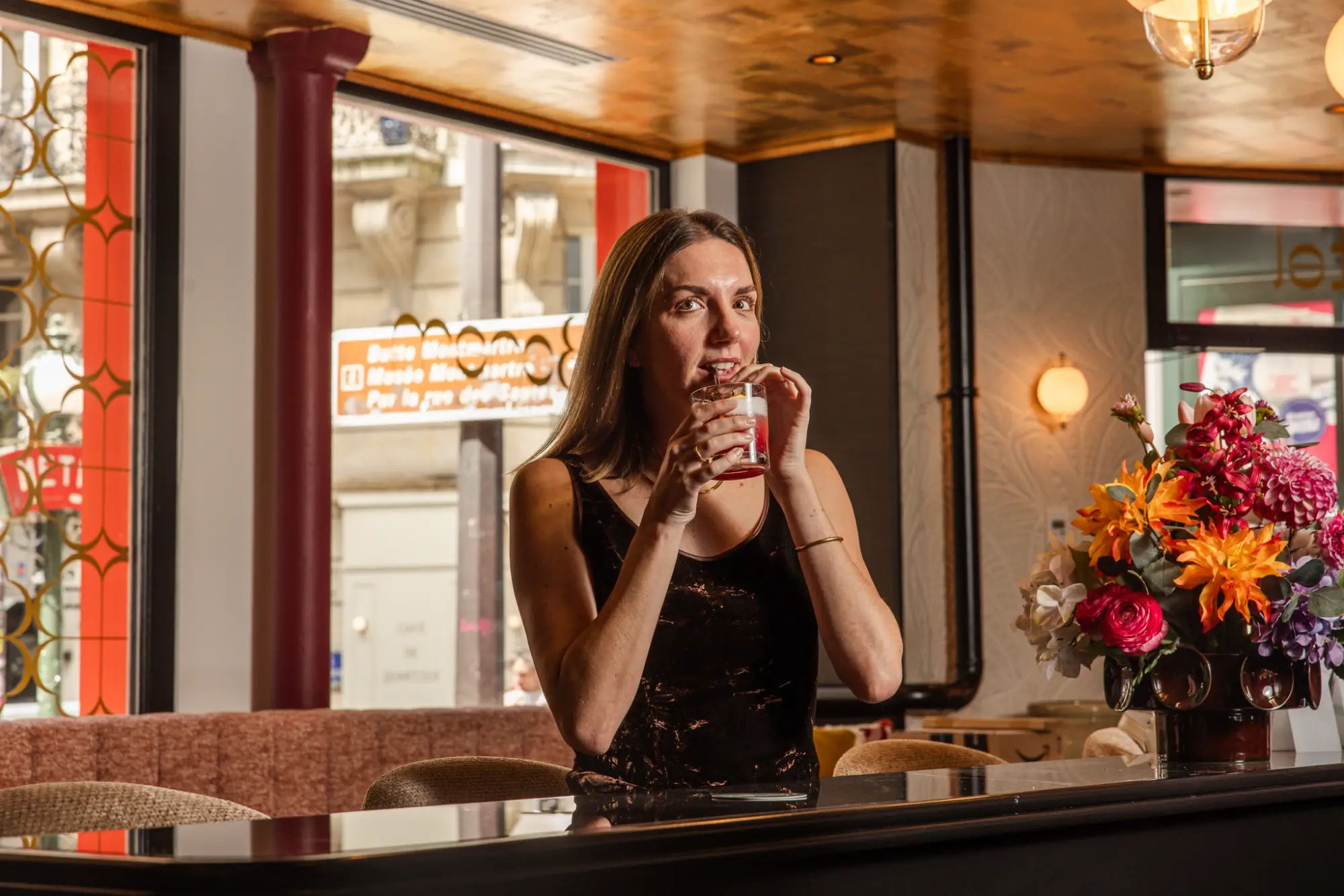 Une femme aux cheveux longs, portant un débardeur noir, boit un cocktail rouge avec une paille assise dans un bar au mobilier et décoration élégants avec un grand bouquet de fleurs colorées sur le comptoir.