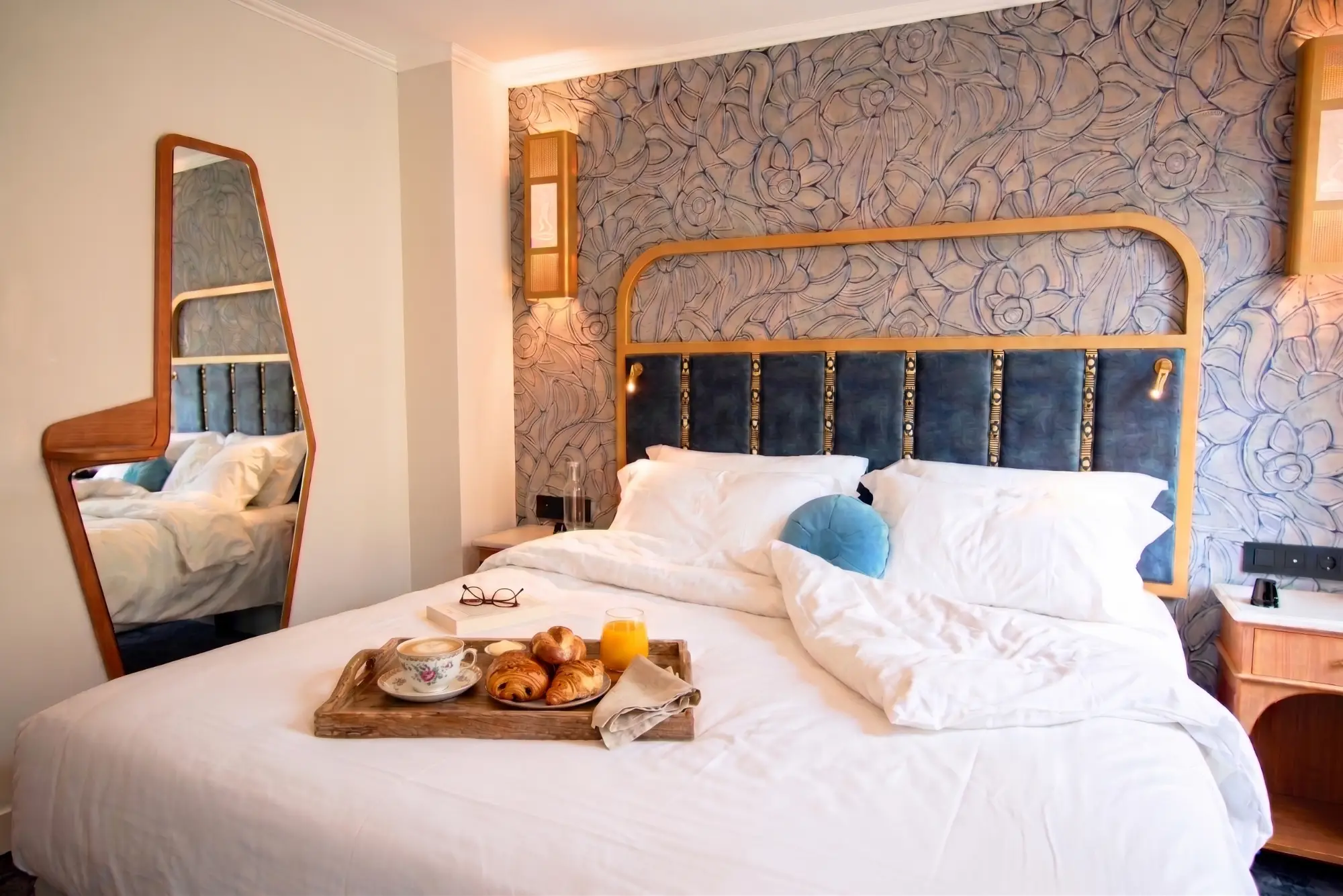 Chambre avec un grand lit blanc, une tête de lit bleue et en bois, un miroir en bois sur le mur gauche, et un plateau de petit-déjeuner avec des croissants, une tasse de café et un verre de jus d'orange posé sur le lit.