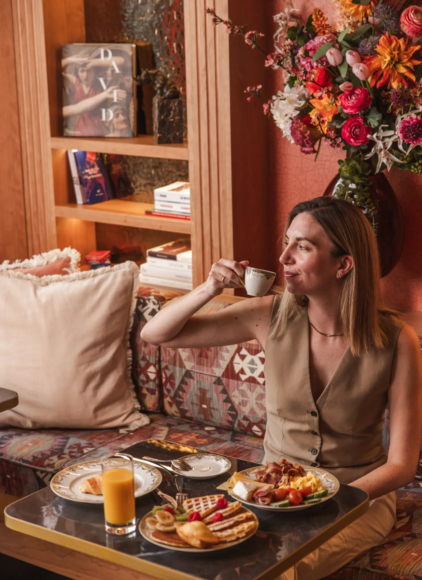 Une femme assise dans un coin salon chaleureux avec coussins et bouquet de fleurs colorées, dégustant une tasse d'espresso, avec un petit déjeuner composé de jus d'orange, gaufres garnies de fruits, croissant, omelette, légumes et charcuterie sur une table devant elle.