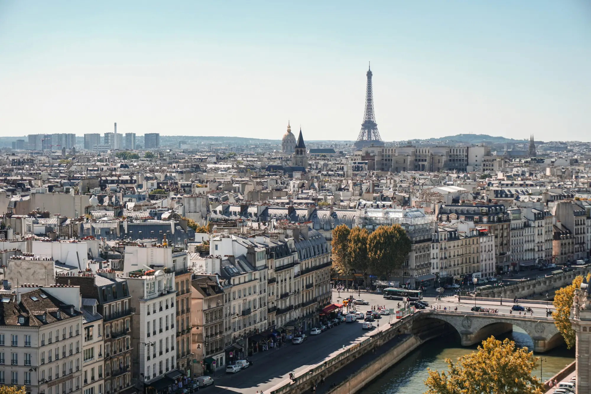 Vue panoramique de Paris montrant la Seine avec un pont et des bâtiments haussmanniens au premier plan, la Tour Eiffel et les toits de la ville en arrière-plan sous un ciel clair.