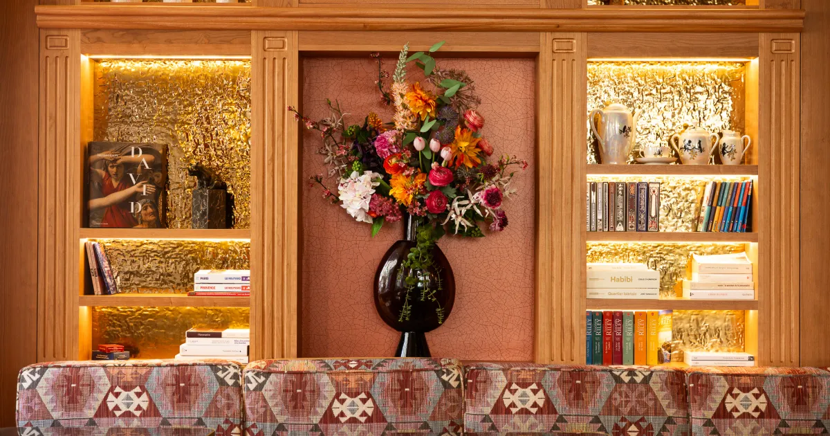 Une étagère en bois avec des étagères éclairées contenant des livres, des vases et des objets décoratifs, avec un grand vase noir central rempli d'un bouquet coloré de fleurs devant un mur texturé, et un canapé couvert d'un tissu à motifs géométriques en premier plan.