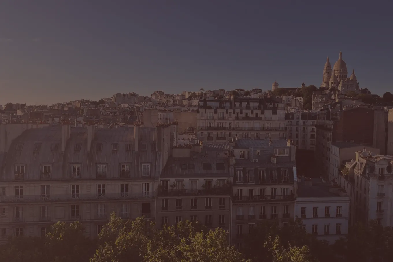 Vue panoramique sur les toits des immeubles parisiens sous un ciel bleu, avec la basilique du Sacré-Cœur visible sur la colline de Montmartre à droite.