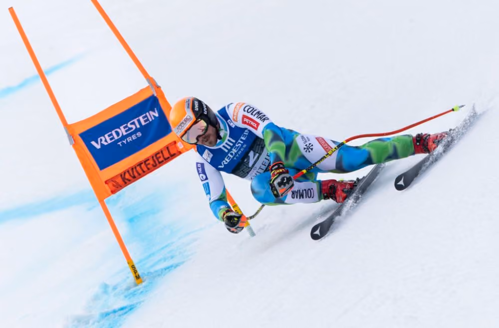 Alpski smučar v oranžni čeladi v zavoju mimo oranžnih vratc med smukom