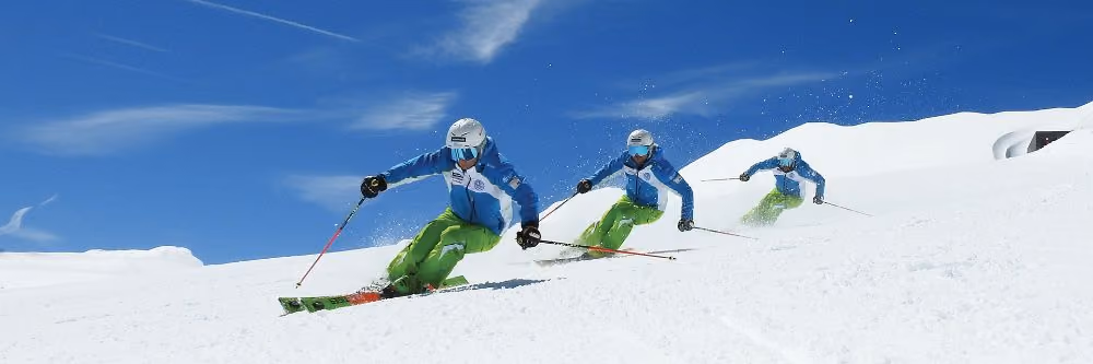 Trije smučarji v vzporednih zavojih na sončni smučarski progi pod modrim nebom