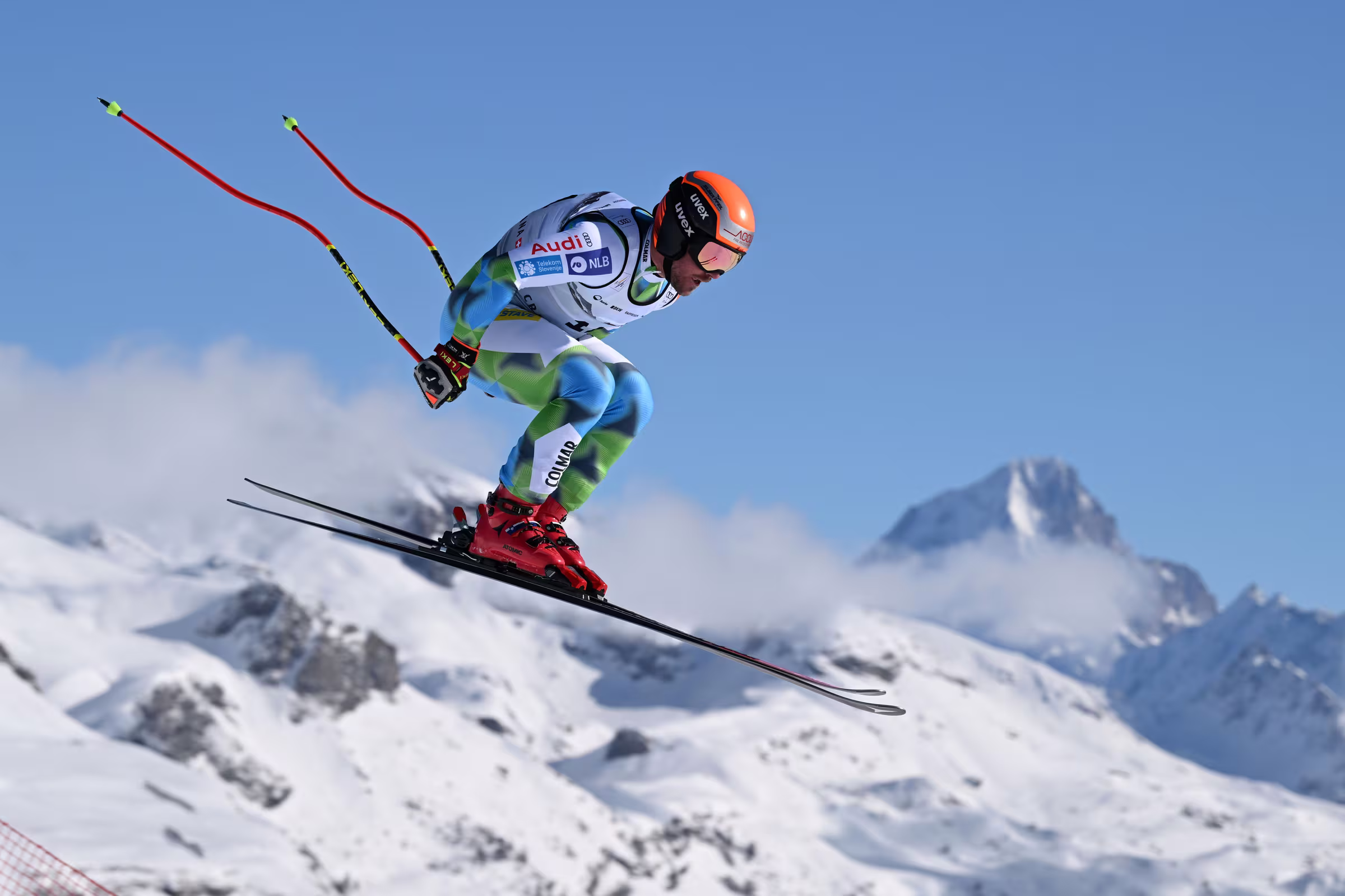 Alpski smučar v skoku čez gorski greben, v ozadju zasneženi vrhovi in modro nebo