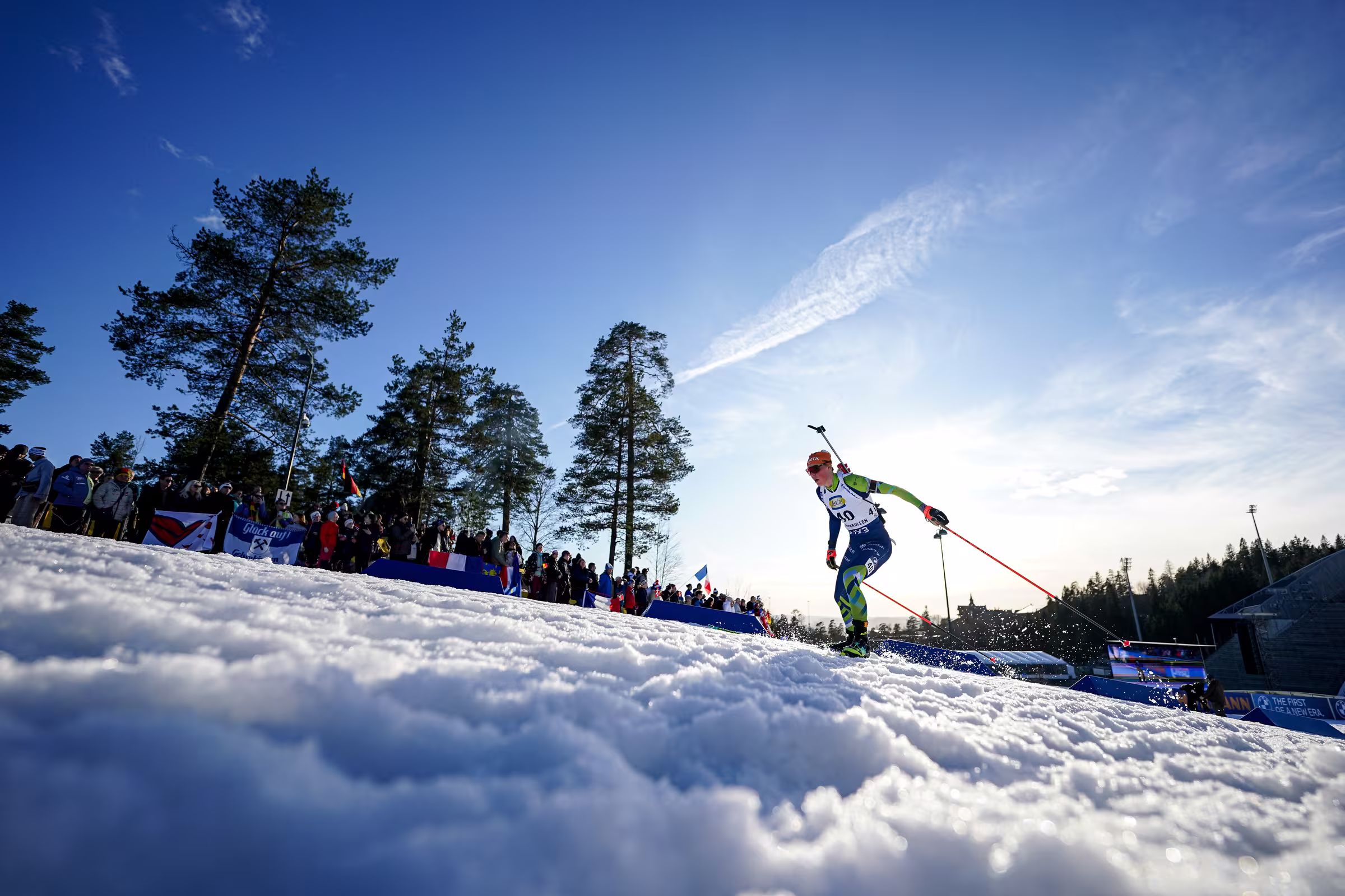 Biatlon na zasneženi progi, ob progi gledalci in borovci pod modrim nebom