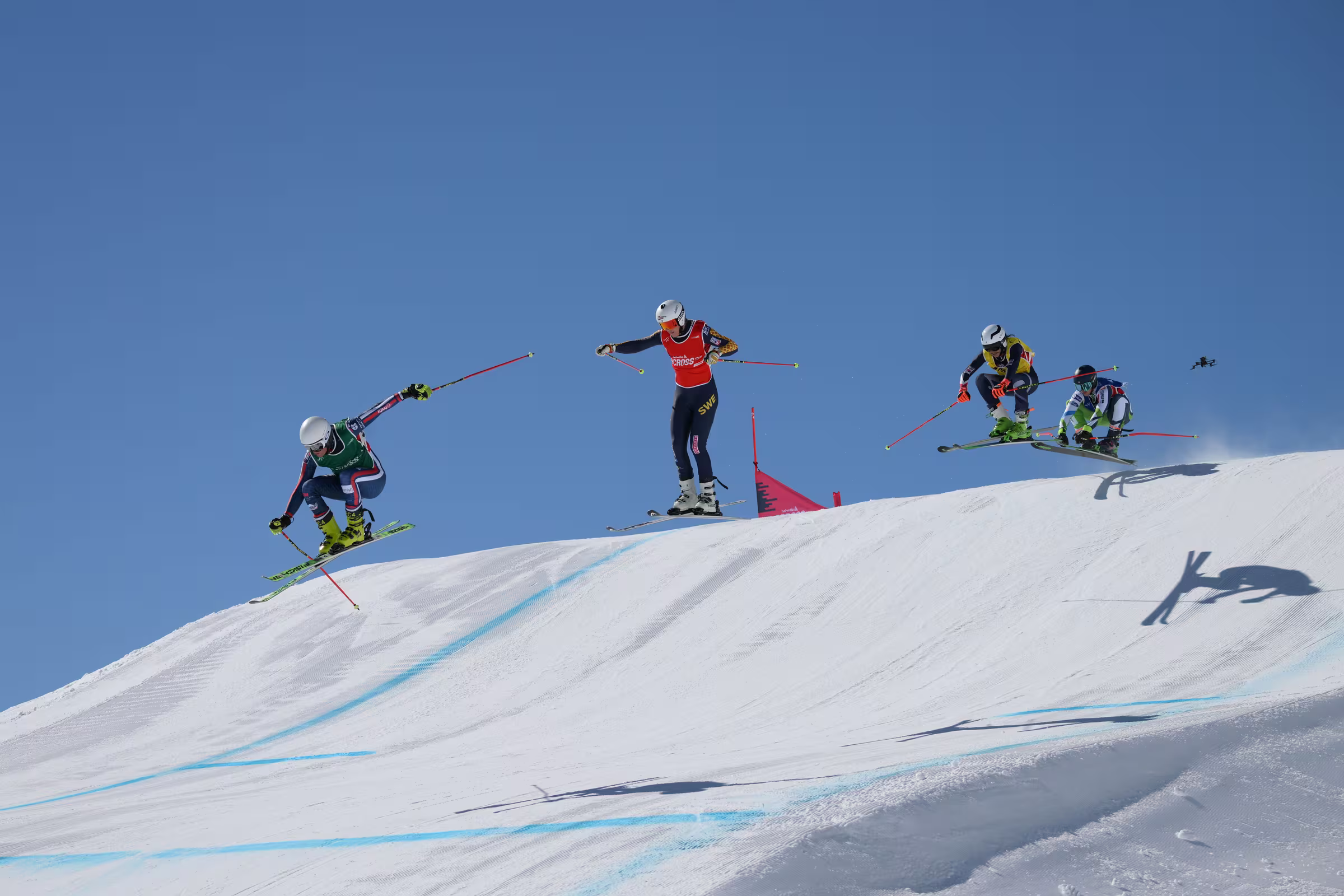 Skicross: smučarji v skoku čez zasnežen hrib pod jasnim modrim nebom
