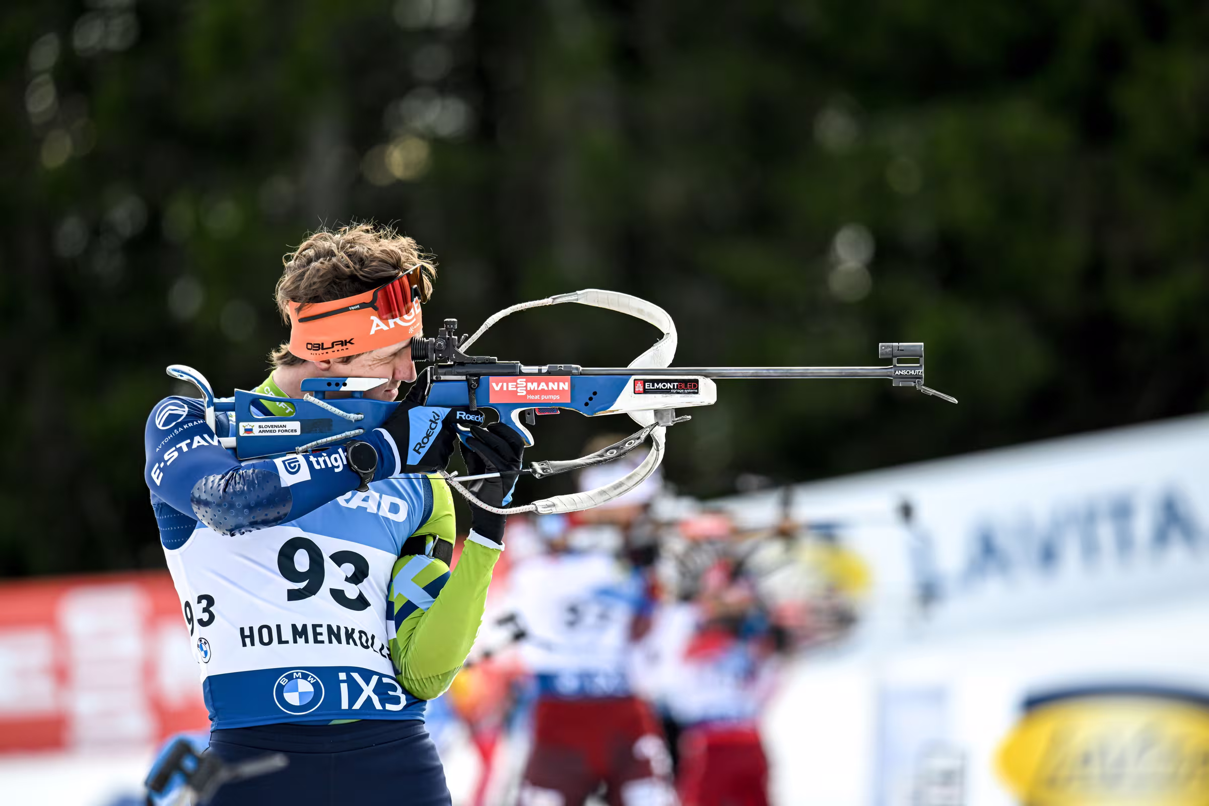 Biatlonec med streljanjem s puško, v ozadju zamegljeni tekmovalci