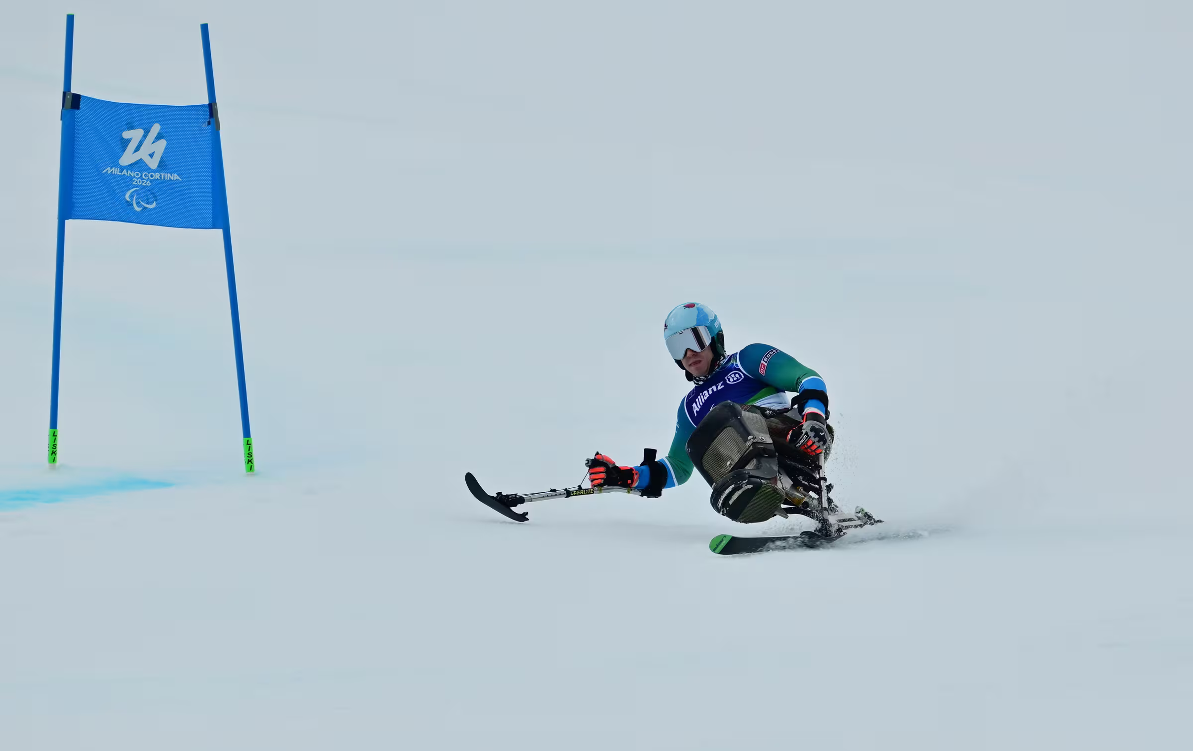 Smučar na sit-skiju v zavoju mimo modrih vratc s paralimpijskim znakom
