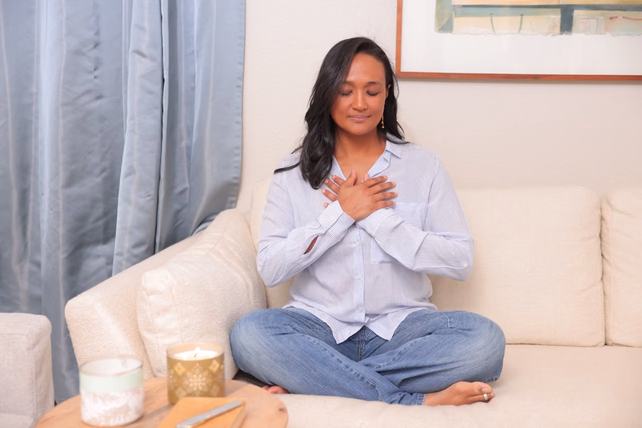 Clementine sits cross-legged on a beige couch with her eyes closed and hands crossed over her chest, appearing calm and meditative.