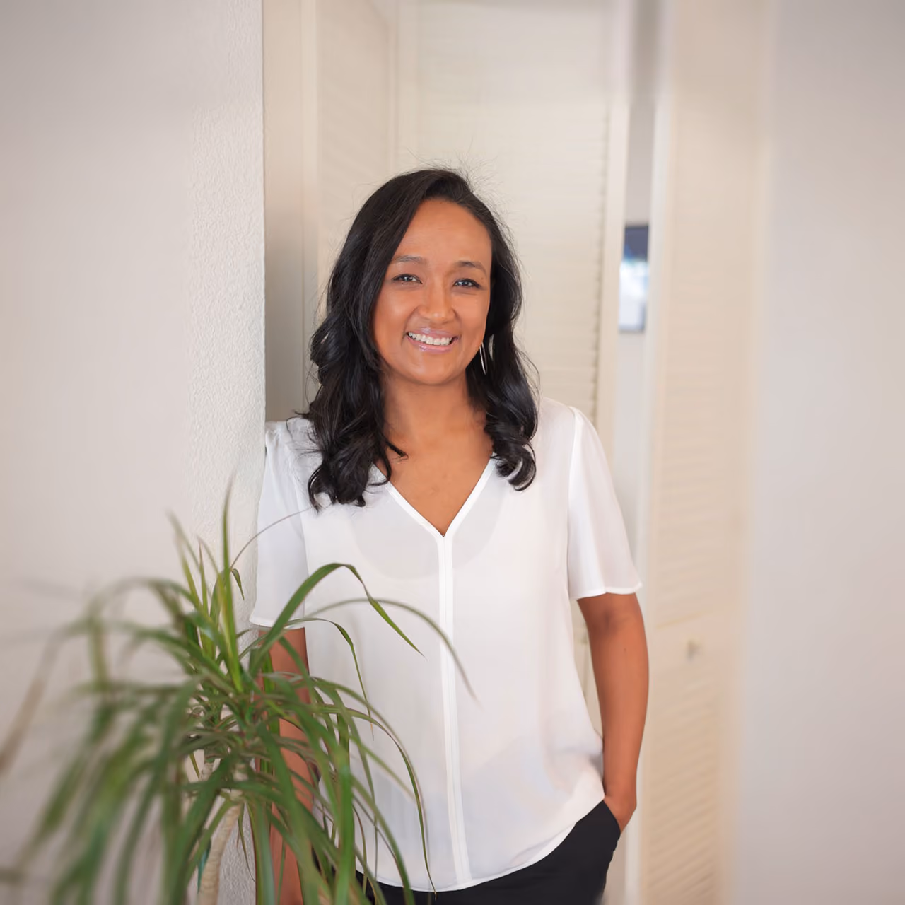 Clementine wearing a white blouse and black pants standing indoors near a potted plant.