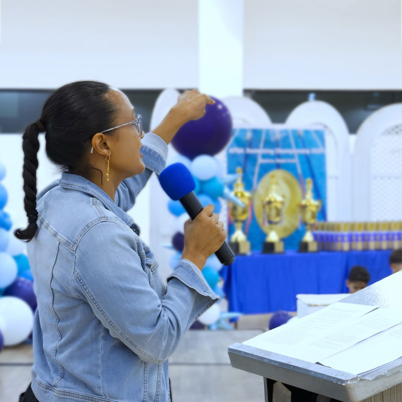 Clementine speaking into a microphone, standing at a podium with papers, in a decorated room with blue and white balloons and trophies in the background.
