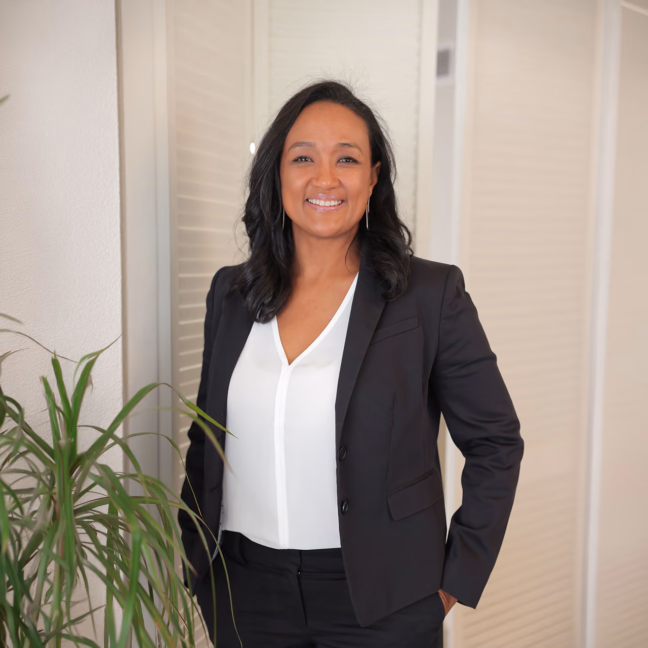 Clementine wearing a black blazer and white blouse standing with one hand in her pocket in a bright indoor space near a green plant.