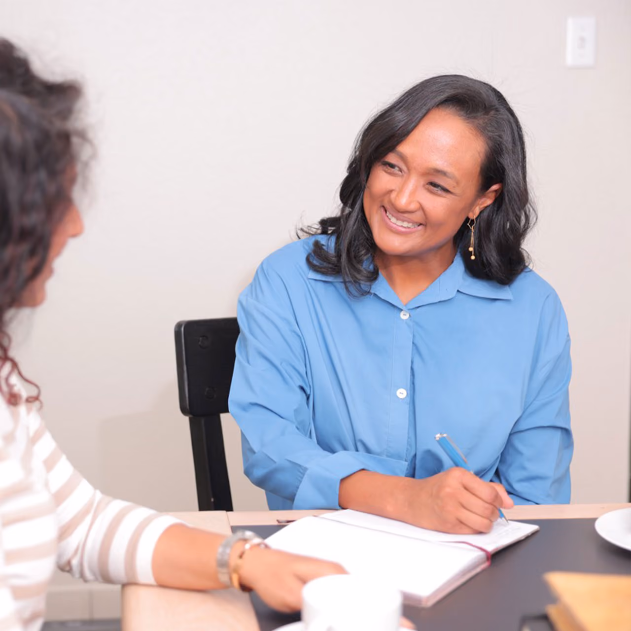 Two women sitting at a table engaged in a friendly conversation, Clementine smiling and holding a pen over an open notebook.